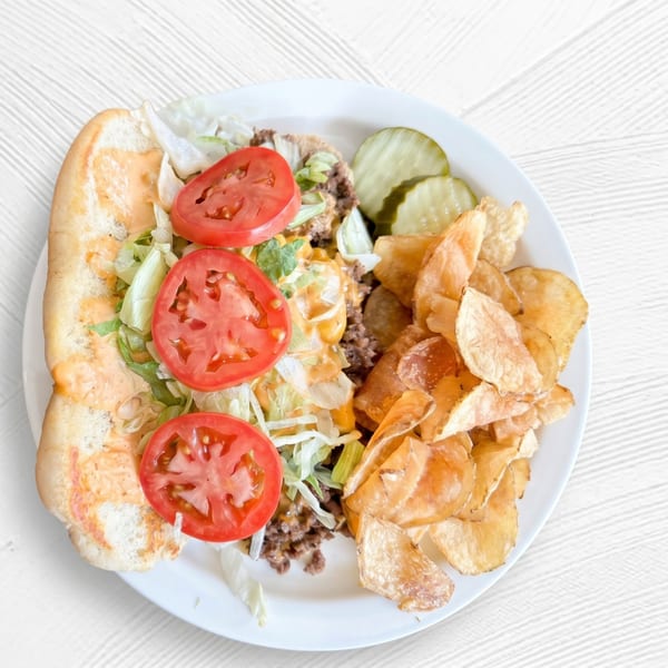 Cheesesteak sandwich with lettuce, tomato, and sauce, served with pickles and potato chips.