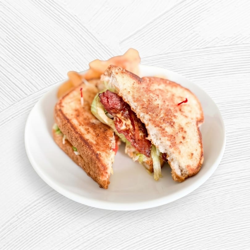 Grilled sandwich halves with bacon, lettuce, and sauce, served with chips on a white plate.