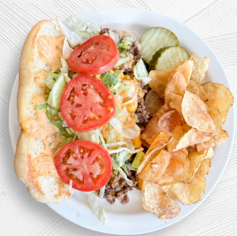 A cheesesteak sub with lettuce, tomato, and sauce, served with pickles and chips on a white plate.