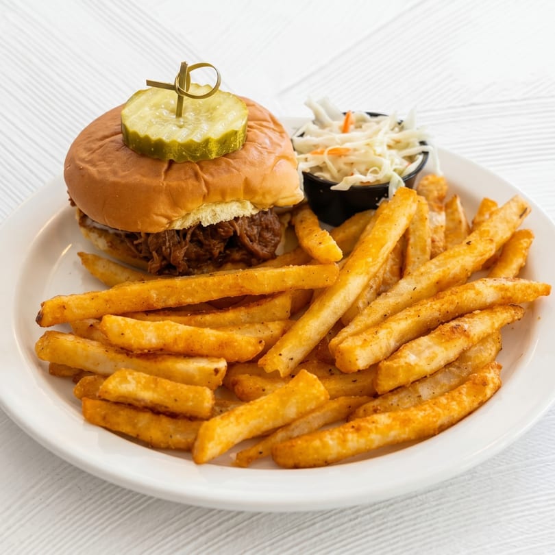 Pulled pork sandwich with a pickle, coleslaw, and a large serving of french fries on a white plate.